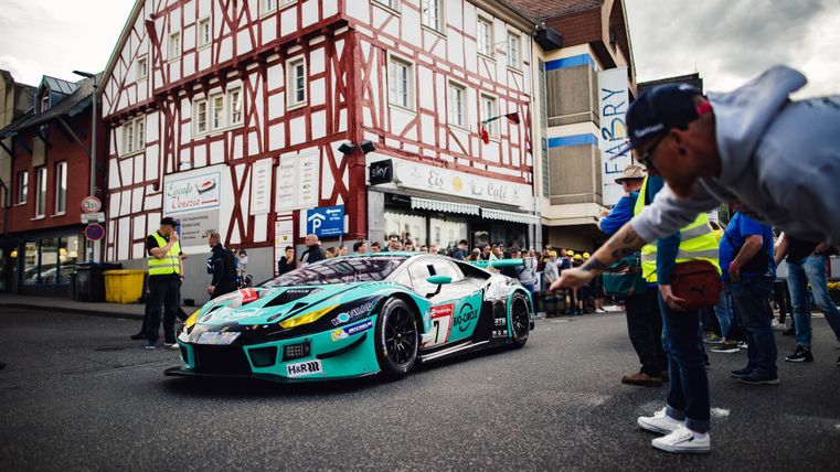 Ein Rennwagen steht auf einer Straße, umgeben von Zuschauern. Im Hintergrund sind traditionelle Gebäude und Sicherheitskräfte zu sehen.