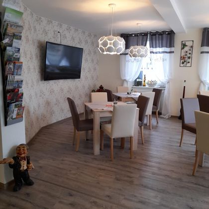 A modern dining room with wooden tables and chairs. There is a television on the wall and windows with daylight.