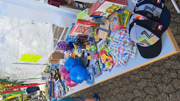 Ein Tisch mit verschiedenen Spielsachen, Hüten und bunten Ballons steht in einem Festzelt. Viele farbenfrohe Artikel sind ordentlich aufgereiht.