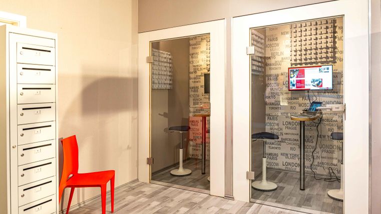 Ein moderner Arbeitsraum mit zwei Glaskabinen und einem roten Stuhl. Im Hintergrund sind Postfächer sichtbar und ein Bildschirm in einer der Kabinen.