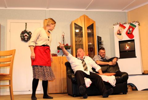 A scene in a living room with three people. A woman stands with an apron while two men sit on a sofa and discuss.