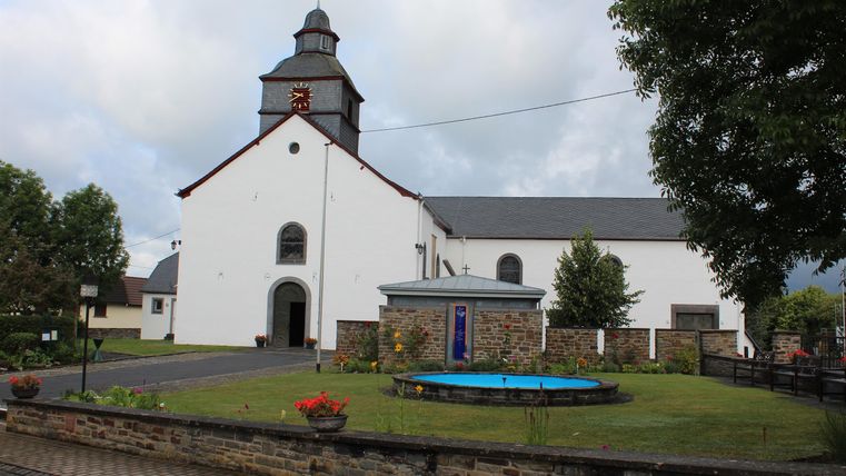 A white church with a tall tower and a small square in front of it. The garden is well-kept and features flowers as well as a small fountain.