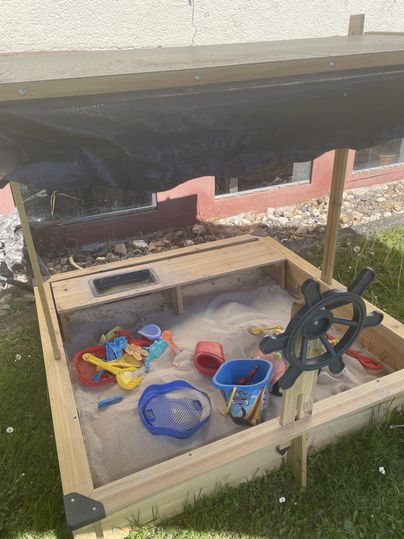 A sandbox with toys and a sun sail. It is made of wood and is located on a green meadow.
