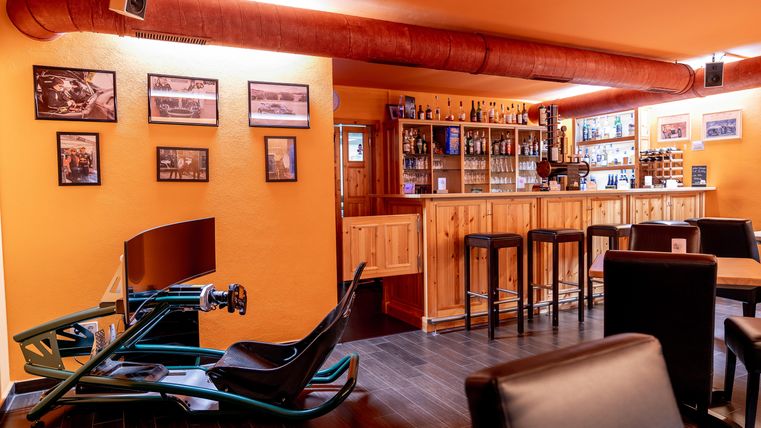 Een gezellige bar met een houten toog en hoge stoelen. Op de voorgrond is een race-simulator te zien die voor vermaak zorgt.