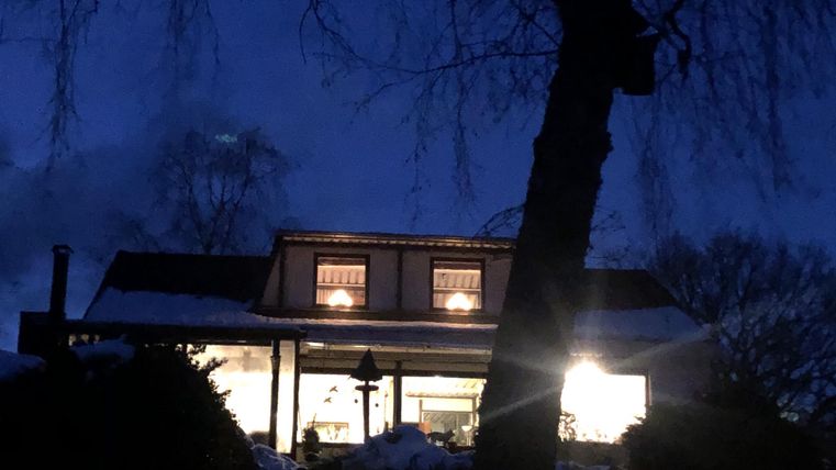 A house at night, illuminated by warm windows. The sky is dark and the surroundings are bordered by trees.