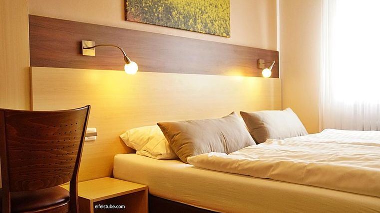 Une chambre moderne avec un lit double et deux coussins. Les murs sont clairs, et une chaise se trouve à côté du lit.