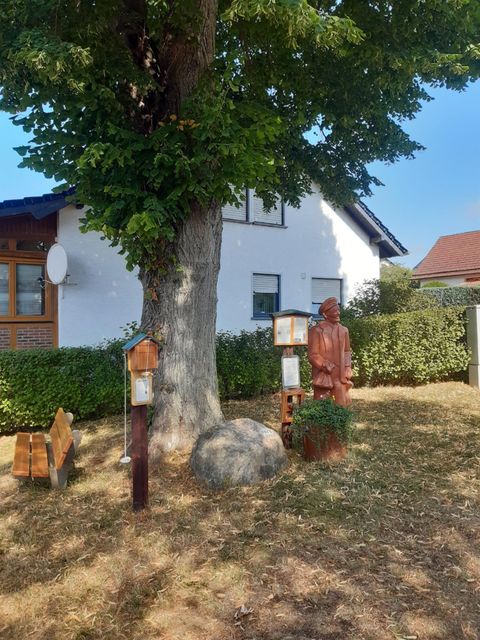 Een verzorgde tuin met een grote boom en een witte huisgevel. Op het gazon staan een houten figuur en twee vogelvoederhuisjes.