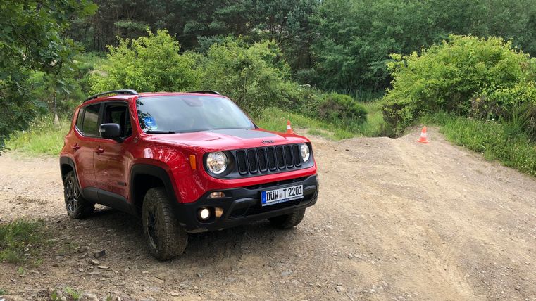 Ein roter SUV steht auf einem unbefestigten Weg in der Natur. Im Hintergrund sind üppige grüne Bäume und Sträucher zu sehen.