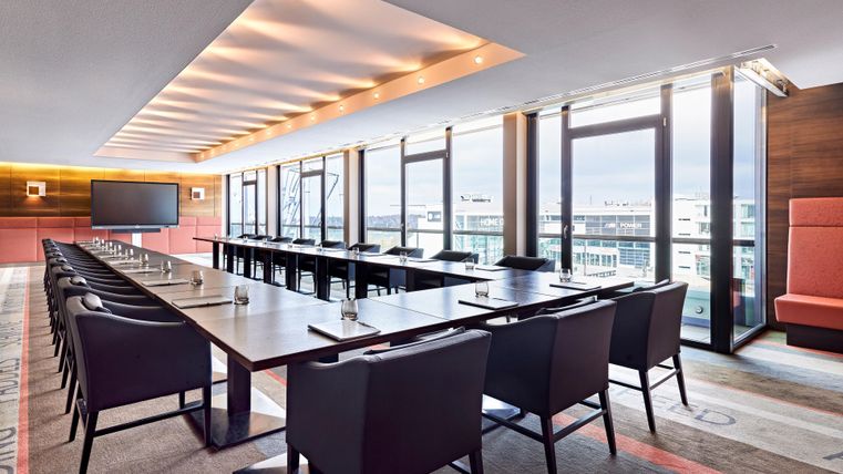 A modern conference room with large windows and a long table arrangement. The room is bright and clean, suitable for meetings or presentations.