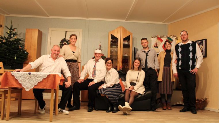 A group of eight people is posing in a festively decorated room. In the background, a Christmas tree and Christmas decorations can be seen.