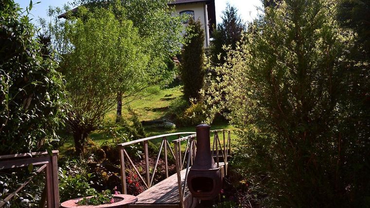 An inviting garden with trees and shrubs. In the background, a wooden walkway and a small grill can be seen.