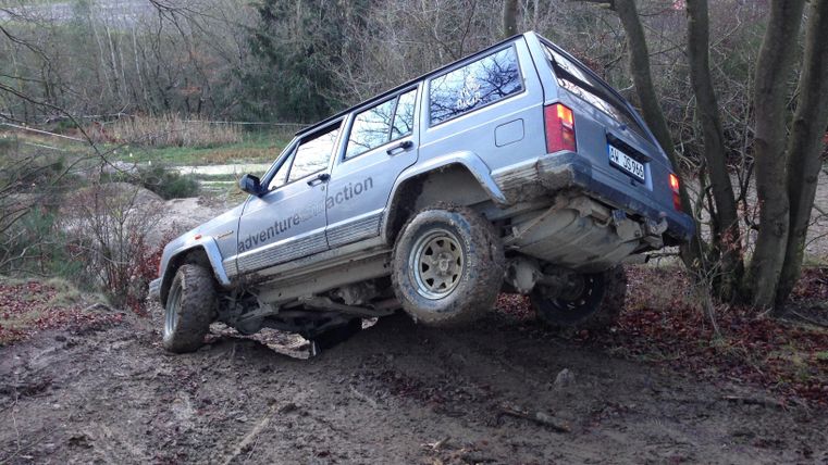 Ein Geländewagen steht schräg auf einem unebenen, schlammigen Untergrund. Umgeben von Bäumen und einer ländlichen Landschaft.