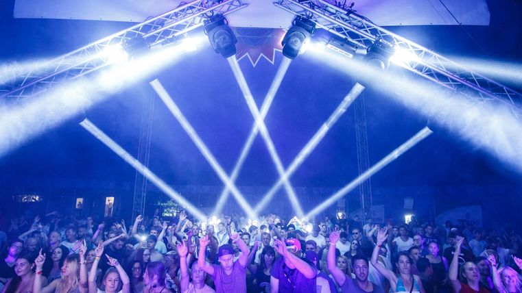 A lively crowd at a concert with colorful light beams. The atmosphere is energetic, and people are dancing enthusiastically.