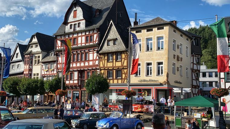 Eine Straße mit historischen Gebäuden und einer farbenfrohen Fassade. Im Vordergrund sind klassische Autos und Menschen zu sehen.