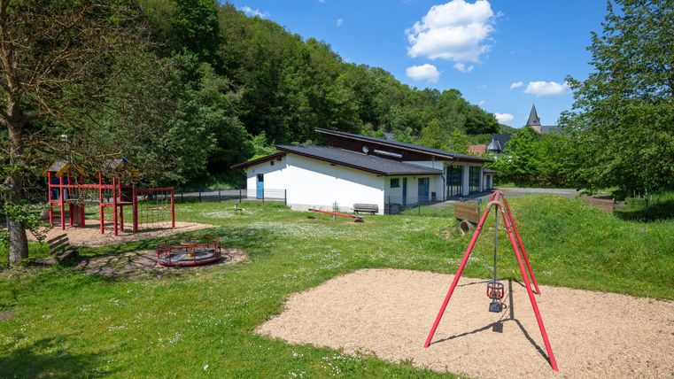 Ein schöner Spielplatz mit einer Schaukel und einer Rutsche. Im Hintergrund ist ein modernes Gebäude und viel Grün zu sehen.