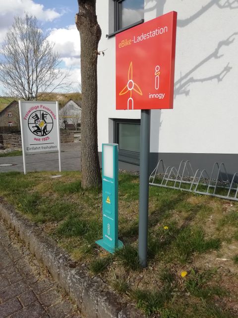 Eine Ladestation für E-Fahrzeuge steht an einem Gebäude. Im Hintergrund ist ein weiteres Schild sichtbar.