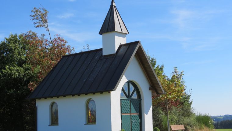 Kleine weiße Kapelle mit grünem Tor und Kreuz auf dem Dach, umgeben von Bäumen und Wiese.