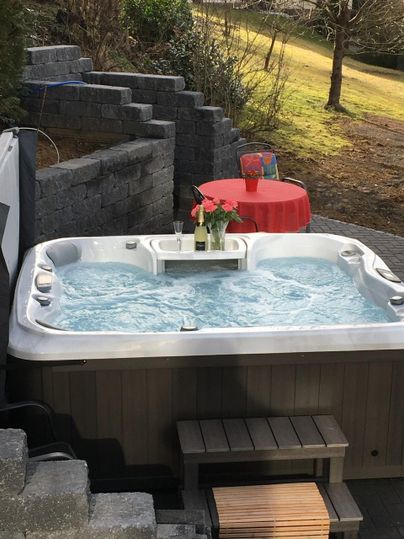 A hot tub outside, surrounded by stone walls and a green meadow. On a red table, there is a bottle of champagne and a floral arrangement.