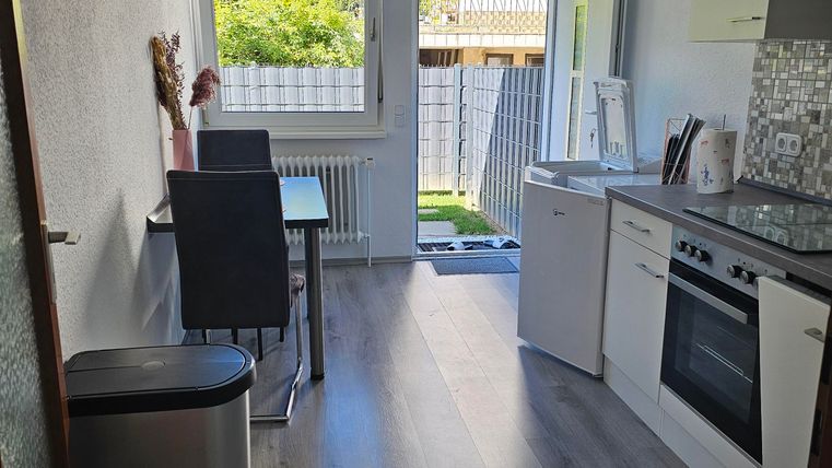 A modern kitchen with a table, chairs, and large windows. The floor has a wood-look finish, and there is access to the garden.