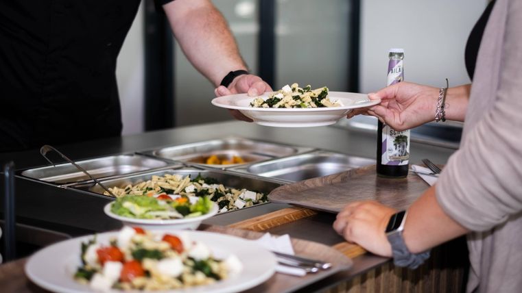 Ein Buffet mit verschiedenen Salaten und Pasta. Eine Person serviert eine Portion auf einem Teller.