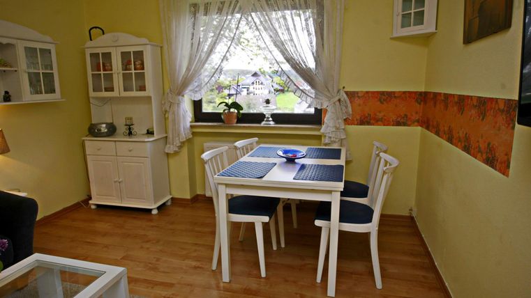 Een gezellige eetruimte met een witte tafel en vier stoelen. De muren zijn geel geschilderd en een raam met gordijnen biedt uitzicht op het buitenzicht.