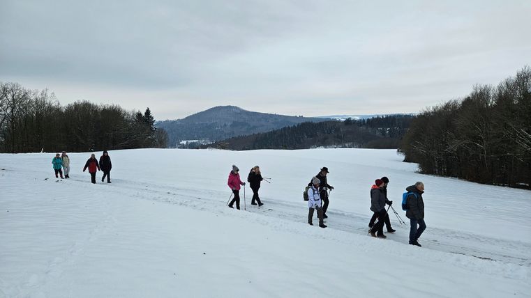 Een groep mensen wandelt door een besneeuwd landschap. Op de achtergrond zijn met sneeuw bedekte heuvels en bosrijke gebieden te zien.