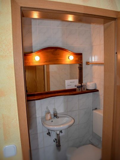 A small bathroom with a sink, a toilet, and a wall mirror. The walls are brightly tiled, and there is a wooden shelf.