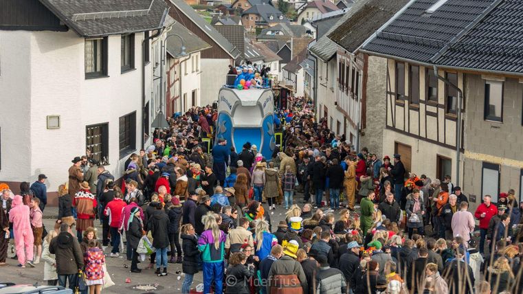 Een grote menigte in kleurrijke kostuums verzamelt zich op een straat. Op de achtergrond is een feestelijk versierde wagen te zien.