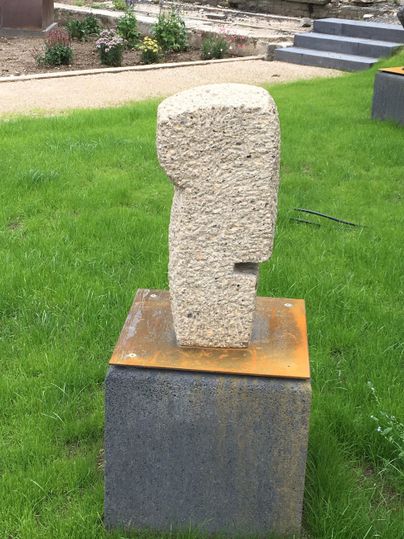 Une sculpture moderne en pierre se dresse sur un socle dans un jardin verdoyant. L'arrière-plan présente des parterres de fleurs soignés et des chemins.
