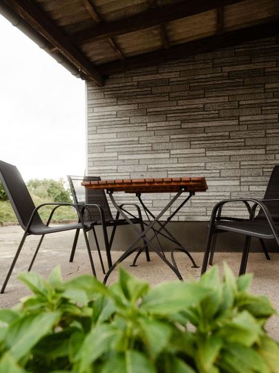 Terrasse mit Tisch und fünf Stühlen, unter einem Dach. Im Vordergrund sind grüne Pflanzen zu sehen.
