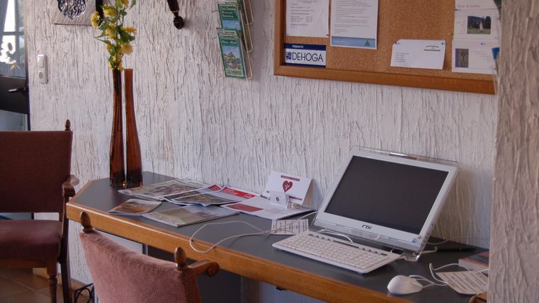 Een kleine bureau met een computer en verschillende informatiematerialen. Op de achtergrond zijn enkele stoelen en een prikbord te zien.
