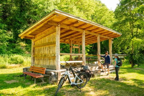Eine Holzunterstand in der Natur mit zwei Radfahrern und einem Fahrrad. Die Umgebung ist grün und sonnig.