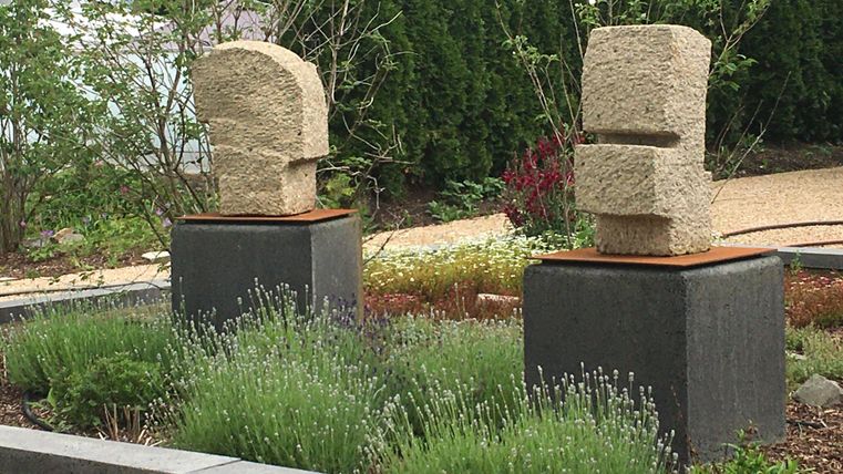 Deux sculptures modernes en pierre se tiennent sur des socles carrés dans un jardin. Entourée de plantes vertes et d'arbustes, la scène crée une atmosphère calme.