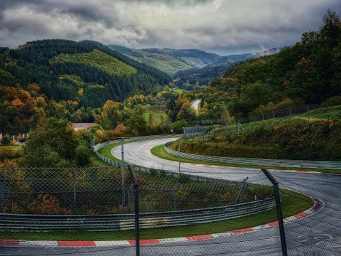 Eine kurvenreiche Rennstrecke umgeben von grünen Hügeln und herbstlichen Bäumen. Der Himmel ist bewölkt und die Landschaft wirkt ruhig.