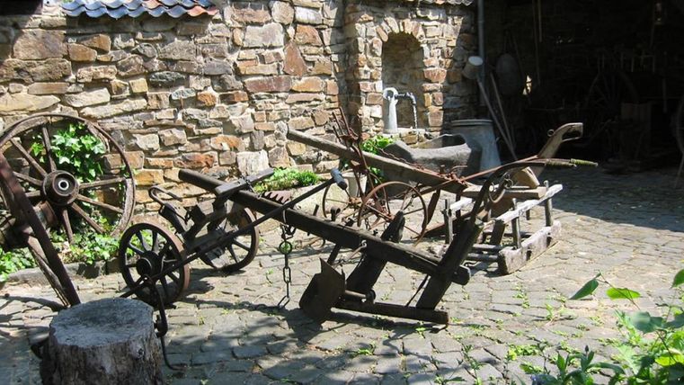 Ein historischer Hof mit alten landwirtschaftlichen Geräten in einem gepflasterten Innenhof. Im Hintergrund sieht man eine steinerne Wand und einige Bäume.