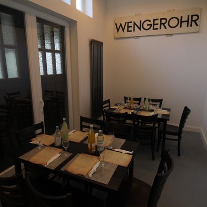 Een restaurant met donkere houten tafels, gedekt met glazen en drankjes. Aan de muur hangt een bord met de tekst 'WENGEROHR'.