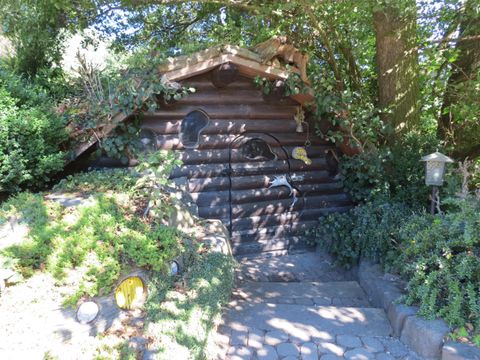 Ein gemütliches Holzhaus, umgeben von grünen Pflanzen. Der Eingang wird von Bäumen und Sträuchern beschattet.