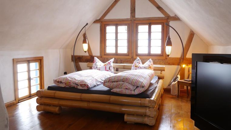 A cozy attic bedroom with a bamboo bed and colorful cushions. Large windows provide plenty of daylight and a pleasant atmosphere.