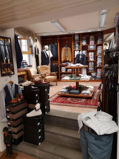 Ein eleganter Bekleidungsladen mit einer vielseitigen Auswahl an Herrenmode. Im Vordergrund sind Schuhe und Kleidung auf Regalen und Tischen präsentiert.
