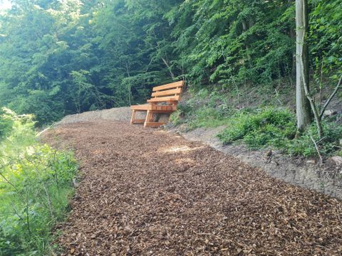Eine gemütliche Bank aus Holz steht auf einem Weg, umgeben von Bäumen und Grün. Der Boden ist mit Holzschnitzeln bedeckt.