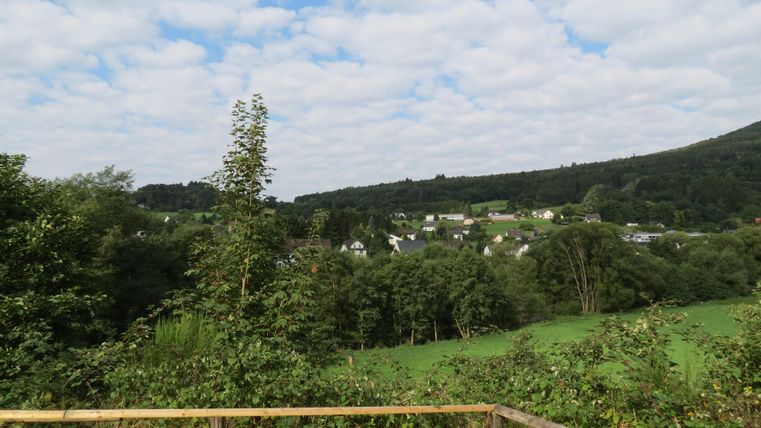 Eine malerische Landschaft mit sanften grünen Hügeln und einer kleinen Siedlung im Hintergrund. Der Himmel ist bewölkt und schafft eine ruhige Atmosphäre.