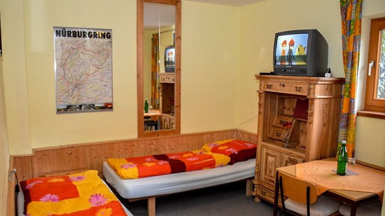 A simple room with two beds, colorful bedspreads, and a small table. On the wall hangs a television and a map of the Nürburgring.
