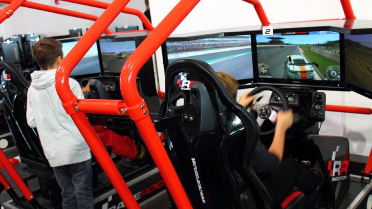 Zwei Kinder spielen an Rennsimulatoren. Die Sitze sind mit roten Rahmen umgeben und die Bildschirme zeigen Rennstrecken.