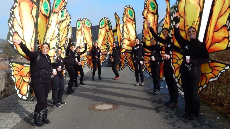 Eine Gruppe von Menschen steht auf einem Weg, trägt große Schmetterlingsflügel und lächelt. Die Szene ist hell und fröhlich, mit einer künstlerischen Atmosphäre.