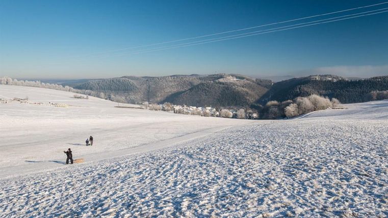 Eine winterliche Landschaft mit Schnee und Bergen im Hintergrund. Zwei Personen genießen den Schnee im Freien.