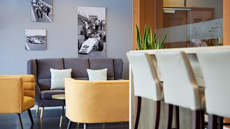 A modern waiting room with comfortable furniture in yellow and gray. Black-and-white photos depicting a racing scene hang on the wall.