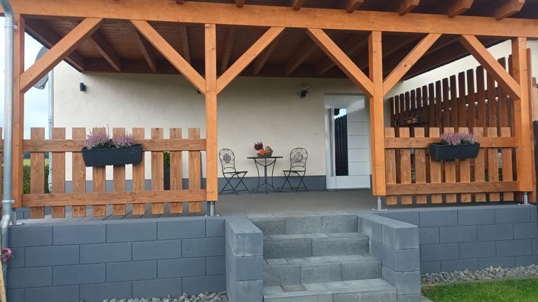 Een gezellige veranda met houten pilaren en een tafel met stoelen. Bloembakken en een goed verzorgd gazon maken het beeld compleet.