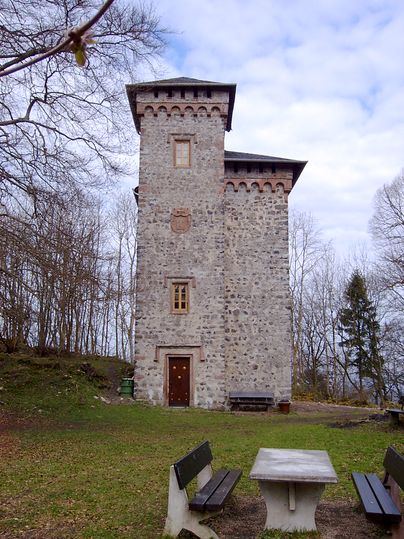 Ein steinerner Turm mit Zinnen und Wappen, umgeben von Bäumen, mit Bänken und einem Tisch im Vordergrund.