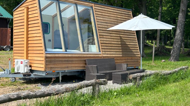 Une maison en bois moderne avec de grandes fenêtres se trouve dans un environnement calme. À côté de la maison, il y a une terrasse avec un parasol et des sièges.