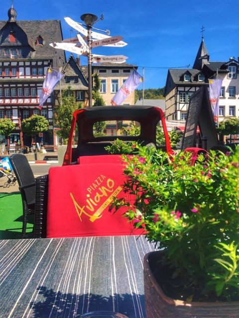 Ein schöner Außenbereich eines Restaurants mit roten Stühlen und einem Blick auf die Umgebung. Im Hintergrund sind historische Gebäude und blaues Himmel zu sehen.